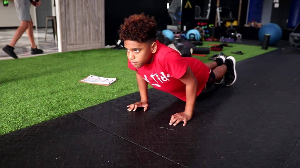 A certified trainer supervising a young athlete performing a bodyweight push up, demonstrating the safest age to start strength training at a gym in New Port Richey, Florida.
